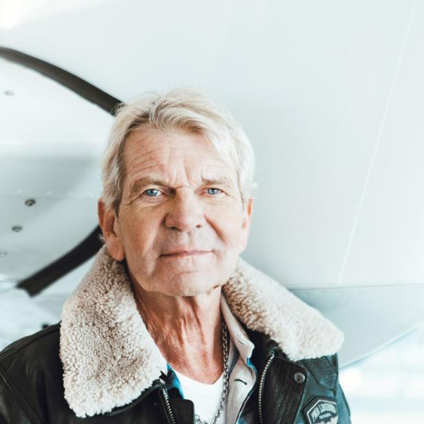 Ein älterer Mann mit grauen Haaren und Fliegerjacke steht vor einem weißen Flugzeug.