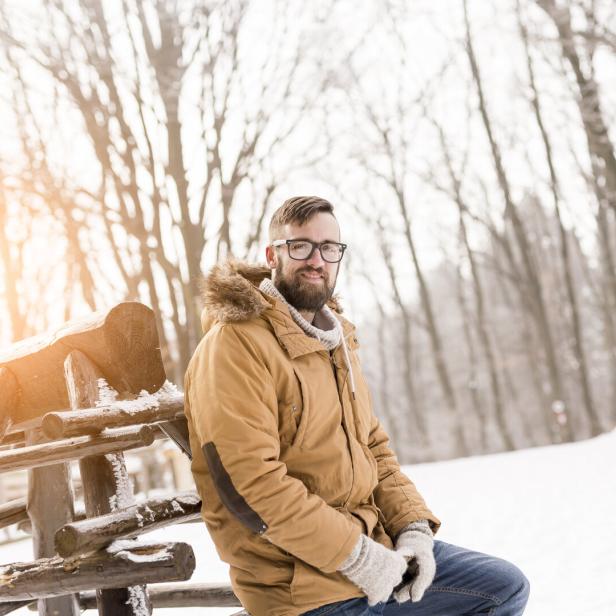 Ein Mann mit Brille und Winterjacke sitzt auf einem Holzzaun im verschneiten Wald.