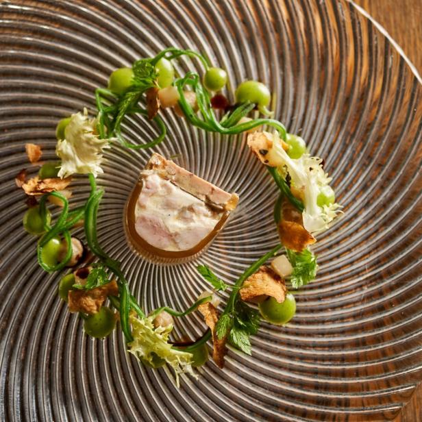 Ein kunstvoll angerichtetes Gourmetgericht mit Terrine, grüner Creme, Kräutern und Salat auf einem gläsernen Teller.