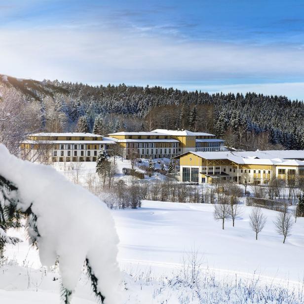 Eine Hotelanlage liegt verschneit vor einem dunklen Wald unter blauem Himmel.