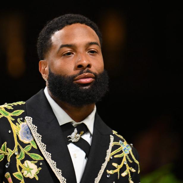 Ein Mann mit Vollbart trägt einen schwarzen Anzug mit floralen Stickereien und Schmuckdetails.