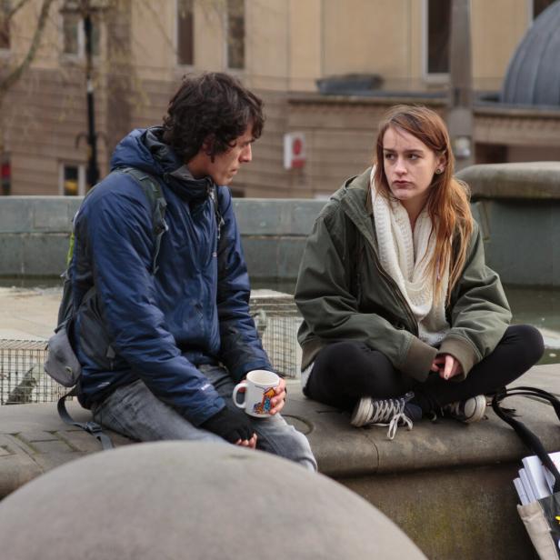 Zwei junge Menschen sitzen auf einer steinernen Umrandung, einer hält eine Tasse, beide wirken nachdenklich.