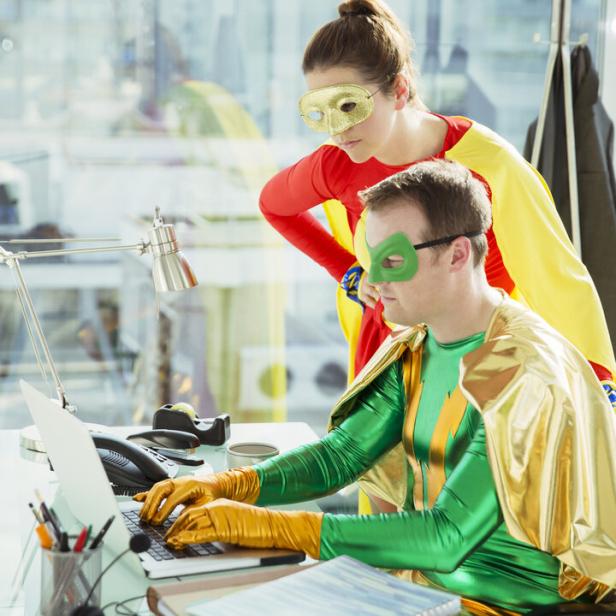 Zwei Erwachsene in Superheldenkostümen arbeiten gemeinsam an einem Schreibtisch mit Laptop in einem modernen Büro.