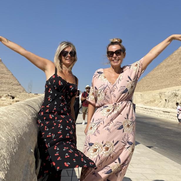 Zwei Frauen posieren vor den Pyramiden von Gizeh mit erhobenen Armen.