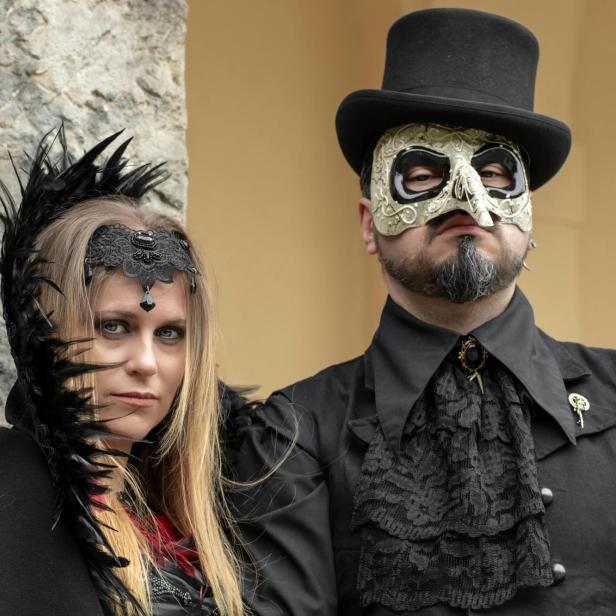 Zwei Personen in opulenter Gothic-Mode mit Spitze, Federn, Maske und Zylinderhut.
