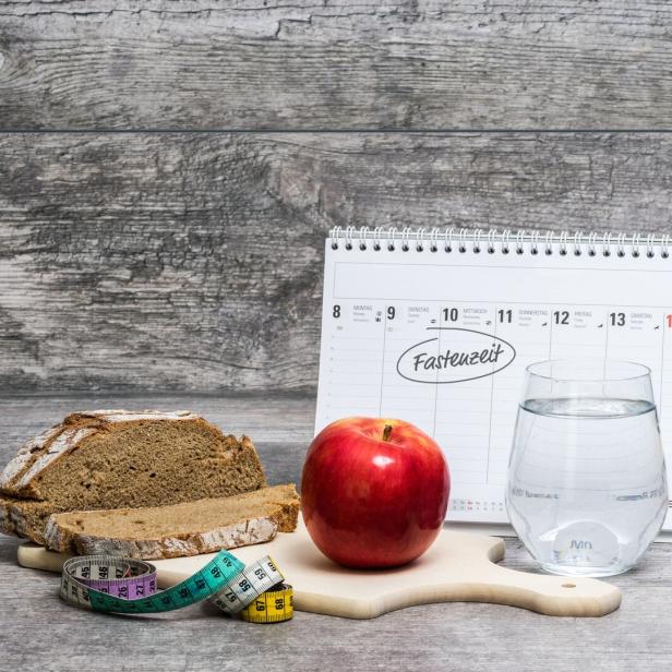 Ein Apfel, ein Glas Wasser, Brot, ein Maßband und ein Kalender mit dem Wort „Fastenzeit“ auf einem Holztisch.