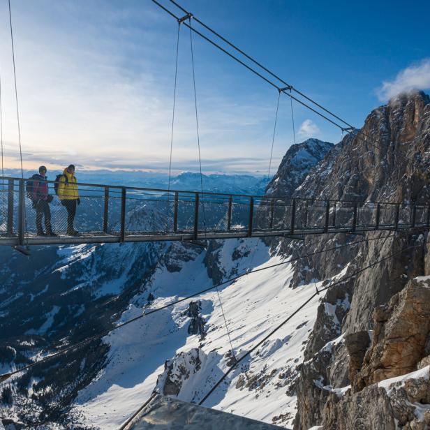 Zwei Personen stehen auf einer Hängebrücke hoch über einer verschneiten Berglandschaft bei Sonnenschein.
