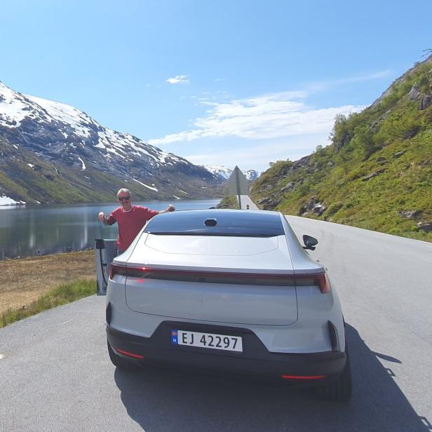 Ein Mann steht neben einem weißen Auto auf einer Straße an einem See, umgeben von Bergen mit Schnee und grüner Vegetation.