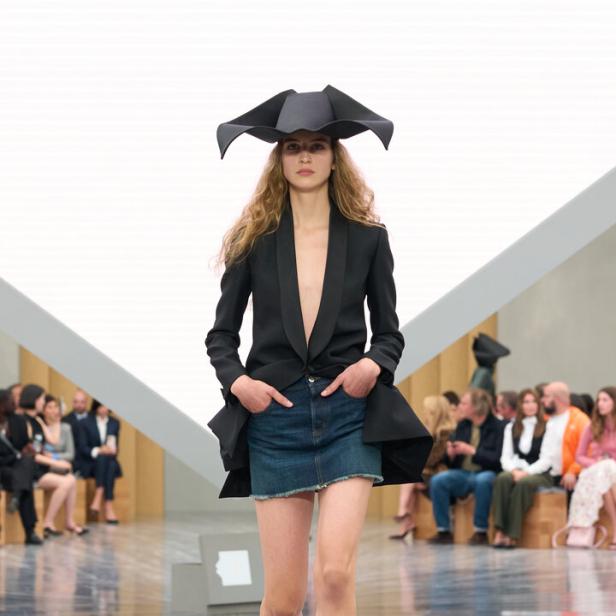 Eine Frau trägt einen tief ausgeschnittenen schwarzen Blazer, einen Jeansrock und einen auffälligen schwarzen Hut auf einem Laufsteg.