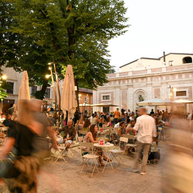 Ein belebter Platz mit vielen Menschen an Tischen unter Sonnenschirmen vor einem Gebäude.