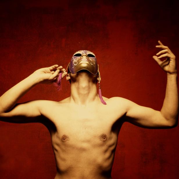 Oberkörper einer Person mit ausgebreiteten Armen und gold-violetter Maske vor rotem Hintergrund.