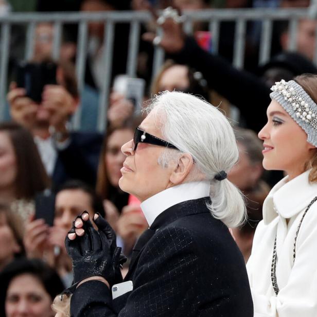 Karl Lagerfeld mit Sonnenbrille und Zopf, neben einem Model mit weißem Mantel, blickt aufmerksam in die Ferne.