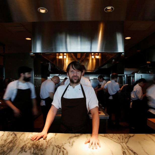 Starkoch und Noma-Chef René Redzepi steht an einer Marmorarbeitsfläche, während mehrere Köche im Hintergrund arbeiten.