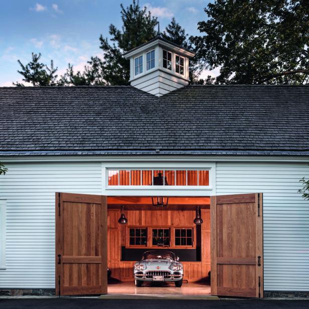 Ein Oldtimer steht in einer offenen, weiß gestrichenen Garage mit Holztoren und gepflegtem Außenbereich.