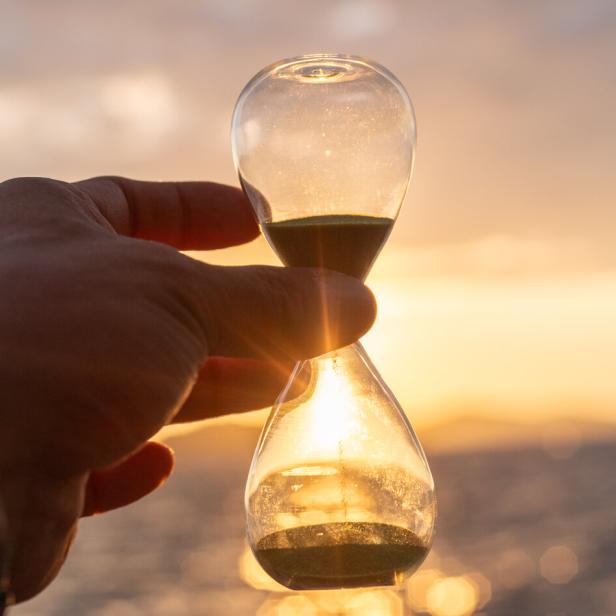 Eine Hand hält eine Sanduhr vor einem sonnigen Himmel.