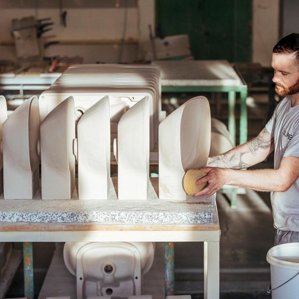 Mann mit kurz geschnittenem Haar und Tattoos schleift mit Schwamm mehrere weiße Keramikwaschbecken auf einem Tisch in einer Werkstatt