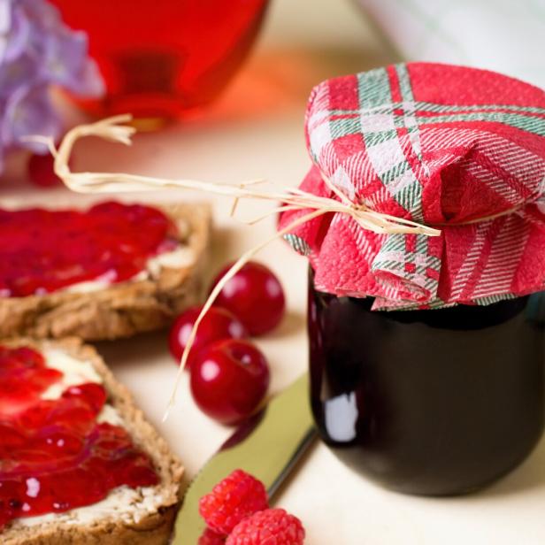 Ein Marmeladenglas mit rot-kariertem Deckel steht neben Kirschen und Brotscheiben mit rotem Aufstrich.