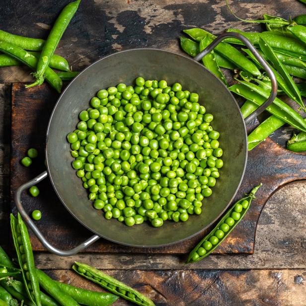 Frische grüne Erbsen liegen in einer Metallschüssel, daneben Schoten, ein Messer und ein Holzbrett auf rustikalem Untergrund.