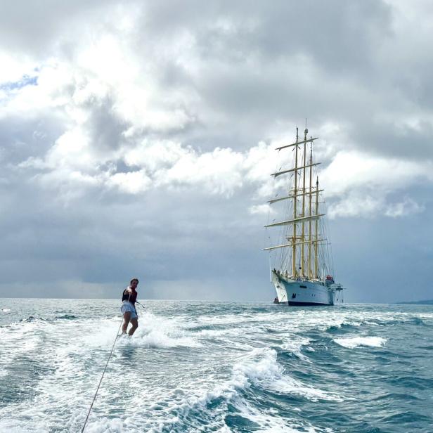 Ein Wasserskiläufer zieht seine Bahnen vor einem großen Segelschiff unter bewölktem Himmel.
