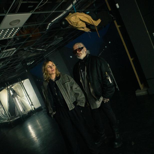 Zwei Personen stehen in einem dunklen Raum mit offenem Deckengebälk, über ihnen hängt eine Schaufensterpuppe kopfüber.