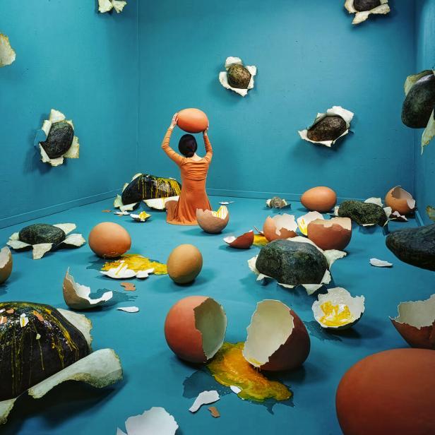 Eine Person in orange sitzt in einem blauen Raum voller zerbrochener Rieseneier und Steine, die aus den Wänden ragen.