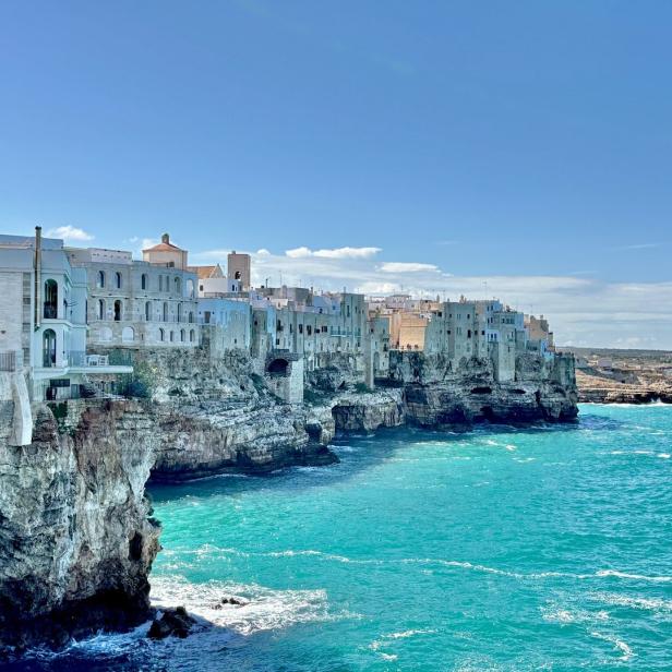 Weiße Häuser thronen auf Klippen über dem türkisfarbenen Meer unter blauem Himmel.