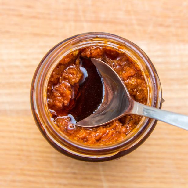 Ein Glas mit rotem Pesto und einem Löffel steht auf einem Holztisch.