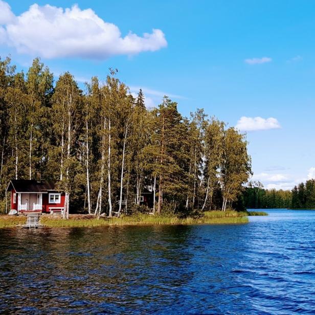 Ein kleines, rotes Haus mit weißen Akzenten steht am Ufer eines Sees, umgeben von Bäumen.