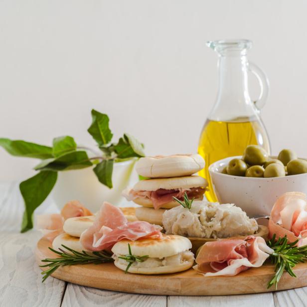 Ein Holzteller präsentiert Oliven, Öl, Brötchen mit Schinken und Kräutern.