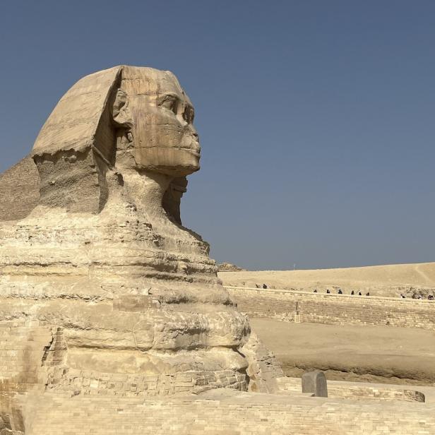 Die Große Sphinx von Gizeh mit Löwenkörper und Menschenkopf steht in einer sandigen Wüstenlandschaft.