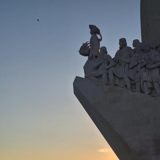 Das Denkmal der Entdeckungen in Lissabon zeigt Skulpturen vor einem Sonnenuntergang über dem Tejo.