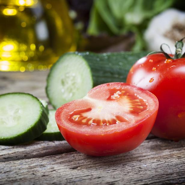 Eine ganze Tomate, eine halbierte Tomate und mehrere Gurkenscheiben liegen auf einem Holztisch.