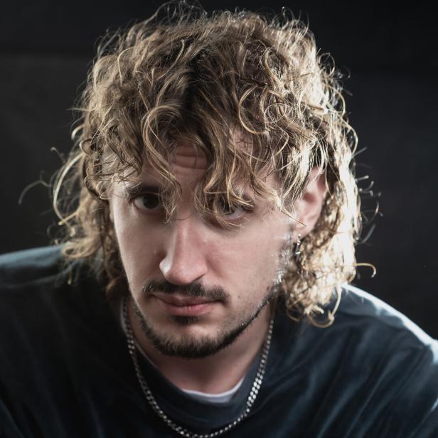 Mann mit lockigen, schulterlangen Haaren, Bart und Kette blickt ernst in die Kamera vor dunklem Hintergrund.