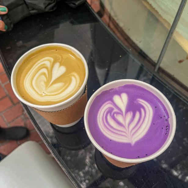 Zwei Becher mit Latte Art, einer mit gelbem und einer mit lila Getränk, stehen nebeneinander auf einem Tisch.
