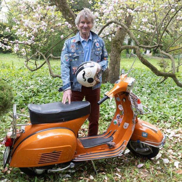 Ein älterer Mann steht im Garten neben einem orangefarbenen Motorroller und hält einen Helm in der Hand.