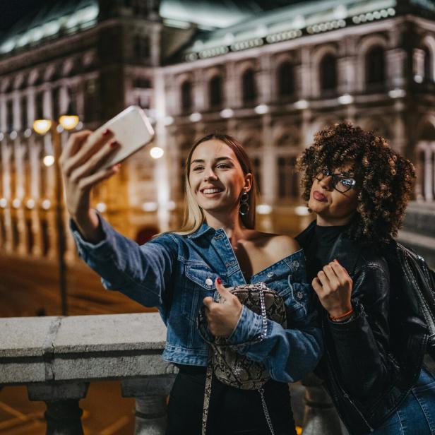 Zwei junge Frauen machen ein Selfie vor einem beleuchteten Gebäude in der Nacht.