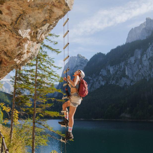 Eine Frau mit Rucksack klettert eine Leiter an einer Felswand über einem See hinauf.