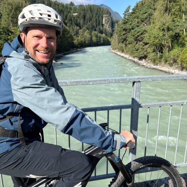 Ein Mann mit Helm und Rucksack fährt auf einem Fahrrad über eine Brücke mit Fluss und Wald im Hintergrund.