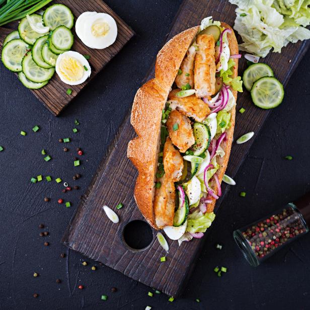 Ein belegtes Baguette mit paniertem Hähnchen, Gurken, Eiern, Salat und Zwiebeln auf einem Holzbrett, daneben frische Zutaten.