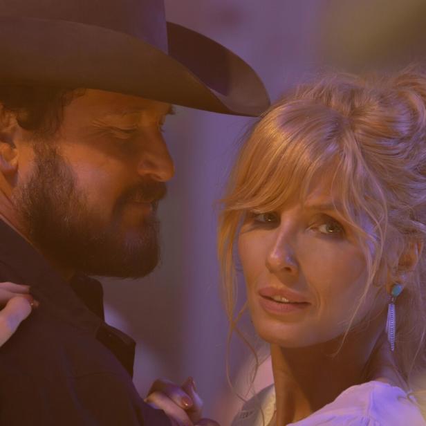 Ein Mann mit Bart und Cowboyhut tanzt eng mit einer blondhaarigen Frau, die nachdenklich in die Ferne blickt.