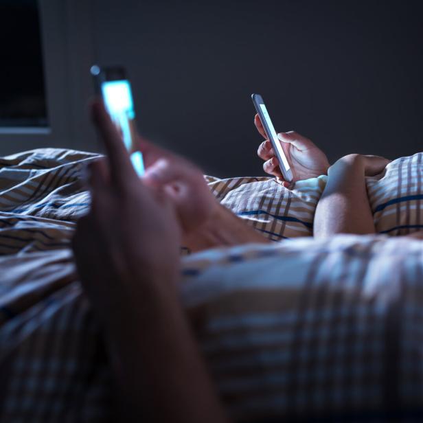 Zwei Personen liegen nebeneinander im Bett und benutzen jeweils ein Smartphone im Dunkeln.
