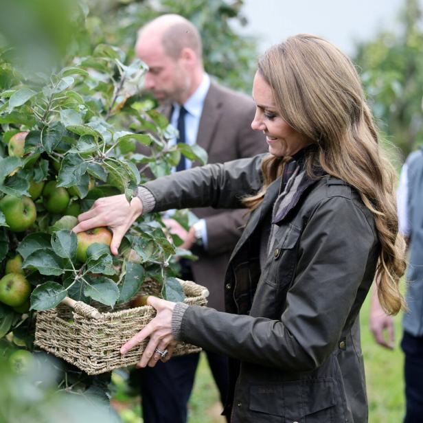 Kate pflückt Äpfel in einen Korb, während William im Hintergrund steht.
