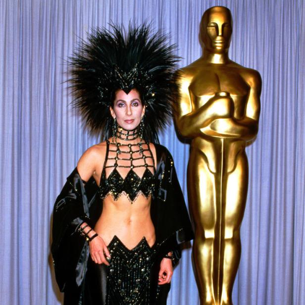 Cher posiert in einem extravaganten, schwarzen Outfit mit Federschmuck neben einer goldenen Oscar-Statue.