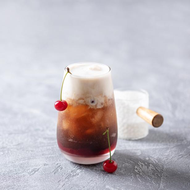 Ein Glas mit einem schaumigen Dirty Soda und Kirschen als Dekoration steht auf einem Tisch.