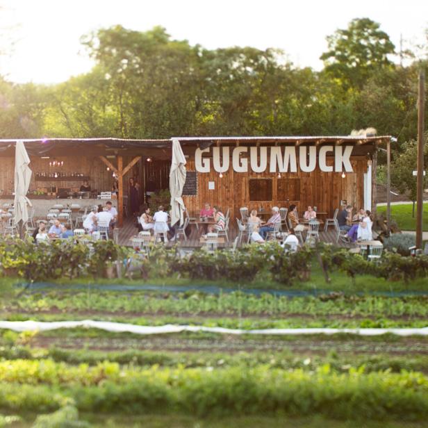 Holzgebäude mit großer Aufschrift „Gugumuck“, davor sitzen Menschen an Tischen im Grünen, links steht eine große Schneckenfigur.