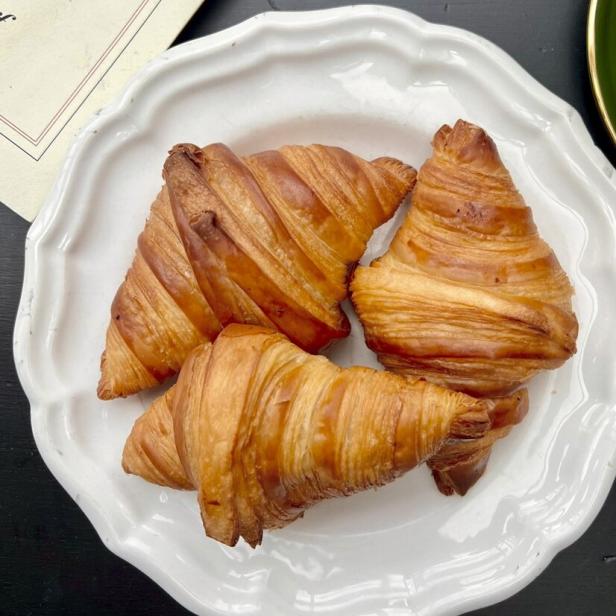 Drei goldbraune, knusprige Croissants liegen auf einem weißen, gewellten Teller vor einem dunklen Hintergrund.