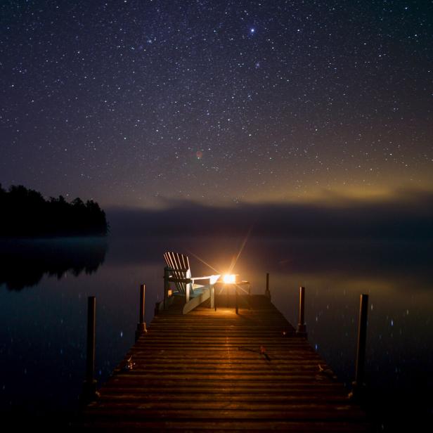 Sternenhimmel über einem stillen See, davor ein beleuchteter Holzsteg mit Stuhl.